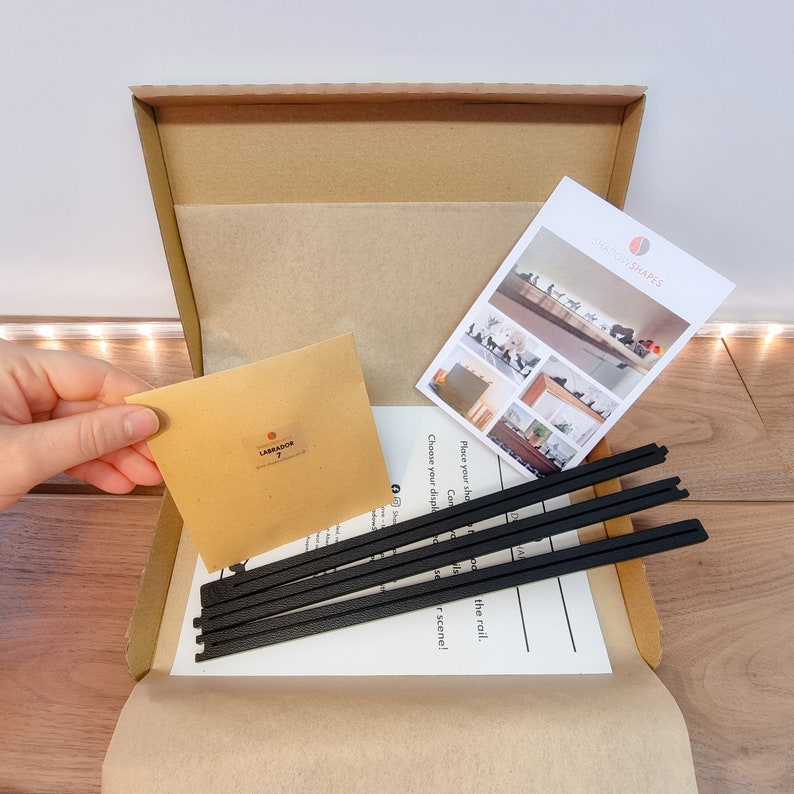 May include: A brown cardboard box with a white paper liner. The box contains black metal rods, a brown paper envelope with the word "LABRADOR" printed on it, and a white card with the words "SHADOWSHAPES" printed on it. The card shows examples of how to use the metal rods to display items on a wall.