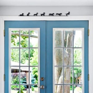 Pode incluir: Uma fila de sete silhuetas pretas de dachshund montadas em uma moldura branca acima de uma porta dupla azul. A porta tem um painel de vidro com vista para uma árvore e folhagem verde.