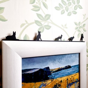 Pode incluir: Uma moldura branca com um passe-partout preto e uma pintura de paisagem em preto e branco dentro. Cinco figuras de cães pretas em silhueta são colocadas no topo da moldura.