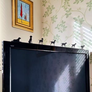Peut inclure: Un téléviseur à écran plat noir avec un cadre argenté est posé sur une table.  Il y a six silhouettes de chiens en métal noir sur le dessus du téléviseur.  Les silhouettes sont disposées en ligne, face au côté droit de l'image.  Le téléviseur est devant un mur avec un motif de papier peint floral vert et blanc.