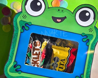 Könnte beinhalten: Eine grüne, froschförmige Geschenkbox aus Pappe mit einem zentralen rechteckigen Sichtfenster, das zwei Süßigkeitenpackungen zeigt. Das Froschdesign hat große schwarz-weiße Augen, einen lächelnden Mund und Schwimmfüße. Der Text NINARTE steht über dem Fenster.