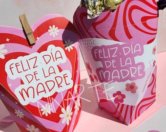 May include: Two pink and red heart-shaped boxes with the text "Feliz dia de la madre" on them. The boxes are decorated with white flowers and swirls. One box is filled with a wrapped chocolate candy.