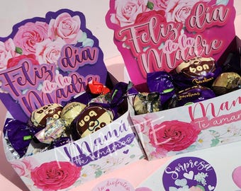 May include: Two pink and purple gift boxes with floral designs and the text "Feliz dia de la Madre" and "Mama te amamos". The boxes are filled with chocolate candies and have pink and white floral stickers.