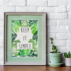 Puede incluir: Una impresión enmarcada con un fondo verde y texto blanco que dice "Keep it simple". La impresión presenta un patrón de hojas verdes. El marco es gris claro y está sobre una mesa de madera.