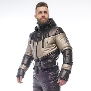 Puede incluir: Un mono de nieve aislado negro y dorado con una chaqueta con capucha y pantalones a juego. La chaqueta presenta un diseño de bloques de color negro y dorado con un cinturón en la cintura. Los pantalones de nieve tienen un aspecto brillante y acolchado.