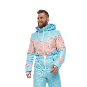 Op de afbeelding: Een lichtblauw en roze skipak met een capuchon en een witte rits. Het pak heeft een riem en is gemaakt van een gewatteerd materiaal.