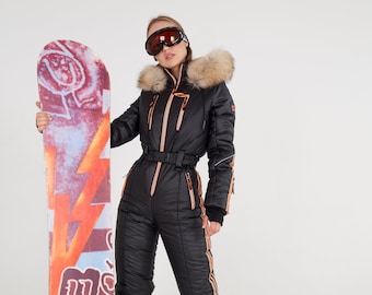 Pode incluir: Uma mulher vestindo um macacão de esqui preto com listras laranja e brancas, um capuz com guarnição de pele e óculos de esqui, segurando um snowboard com um design colorido e o texto "Tecno" nele.