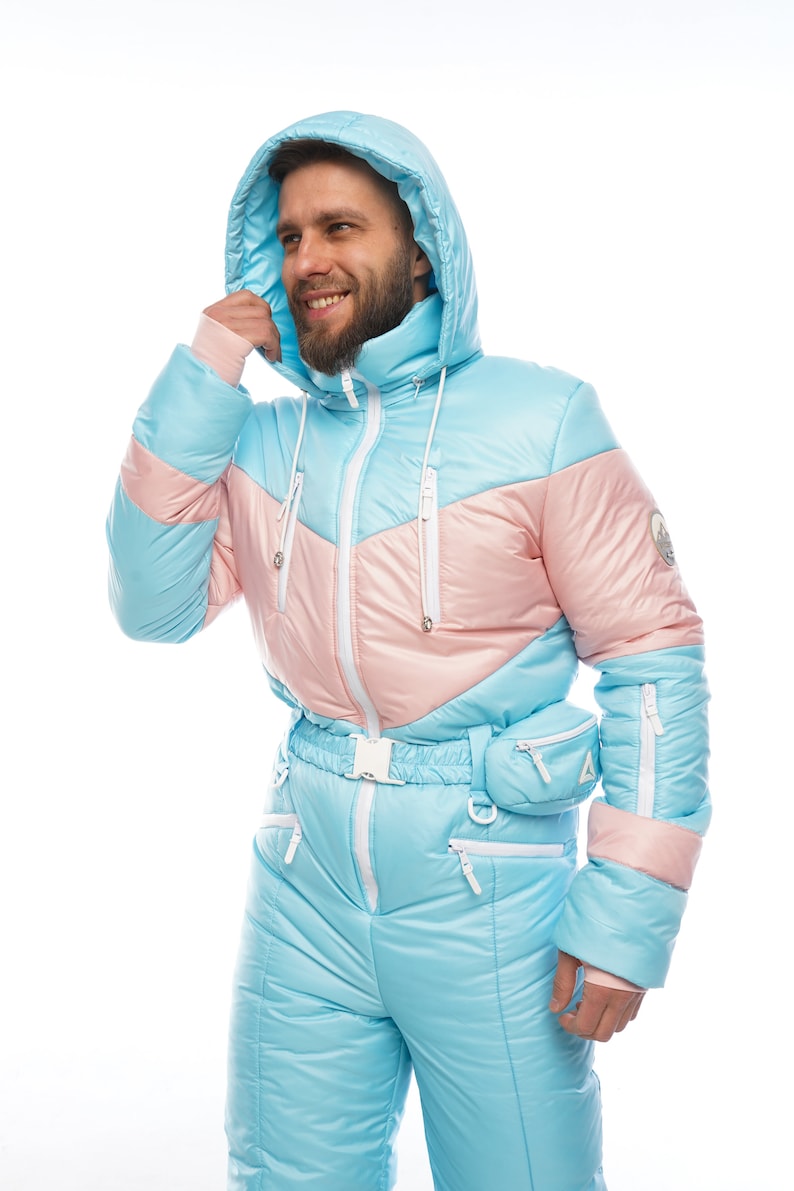 Op de afbeelding: Een lichtblauw skipak met roze en witte accenten. Het pak heeft een capuchon en een ritssluiting. Het pak heeft een riem en zakken.