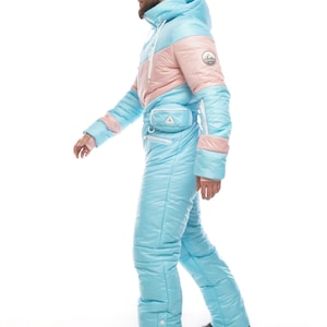 Op de afbeelding: Een lichtblauw en roze skipak met een witte rits en een klein zakje op de heup. Het pak heeft een capuchon en is gemaakt van een pluche materiaal.