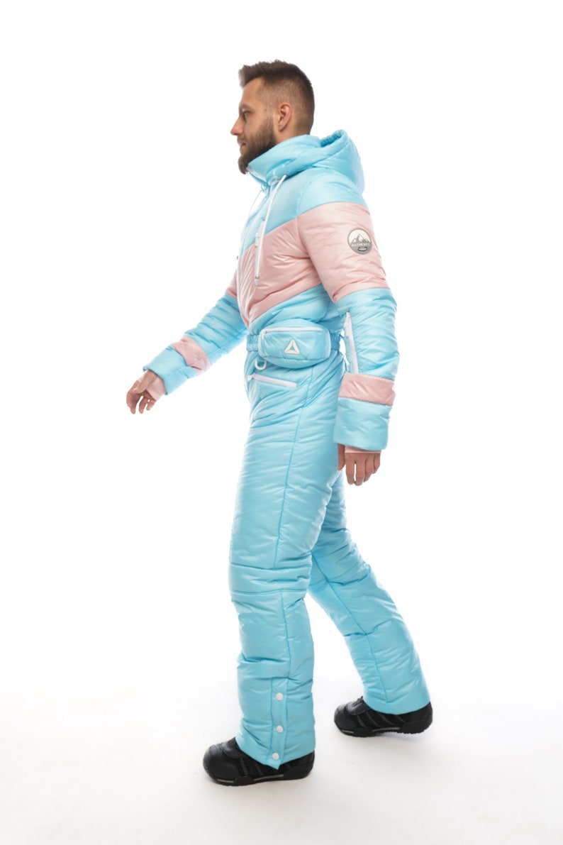 Op de afbeelding: Een lichtblauw en roze skipak met een witte rits en een klein zakje op de heup. Het pak heeft een capuchon en is gemaakt van een pluche materiaal.