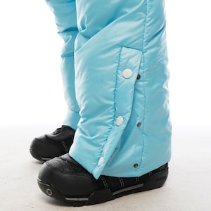 Op de afbeelding: Lichtblauwe sneeuwbroek met witte drukknopen aan de zijkant. De broek is gemaakt van waterafstotend materiaal en heeft een gewatteerd ontwerp.