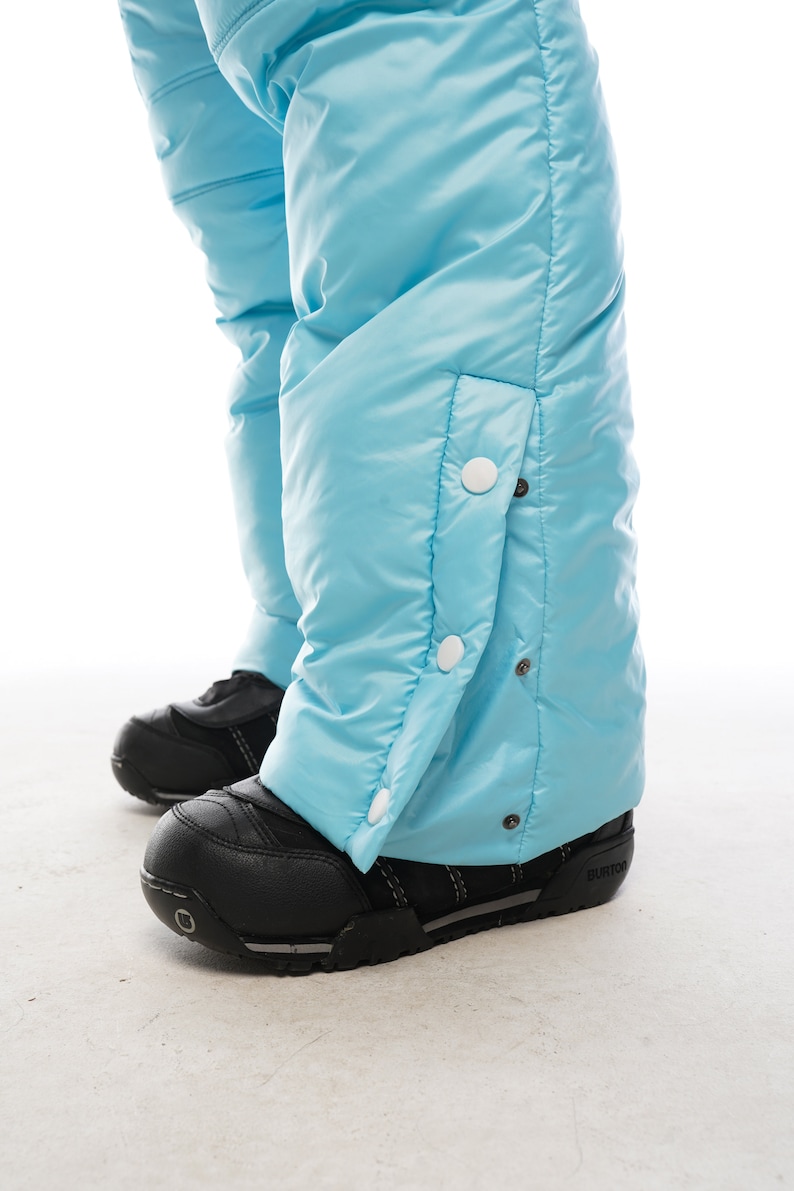 Op de afbeelding: Lichtblauwe sneeuwbroek met witte drukknopen aan de zijkant. De broek is gemaakt van waterafstotend materiaal en heeft een gewatteerd ontwerp.