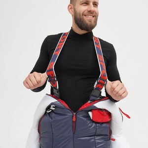 以下が含まれることがあります： 白と赤のアクセントが付いたネイビーのスキーウェアを着た人。ウェアには赤い文字で「SPORT」と書かれたサスペンダーが付いています。