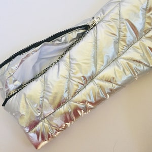 Puede incluir: Bolso de mano plateado acolchado con cremallera negra. El bolso tiene un patr&oacute;n de diamantes y un interior blanco. La cremallera est&aacute; parcialmente abierta, revelando el interior. El material tiene un aspecto brillante y met&aacute;lico.