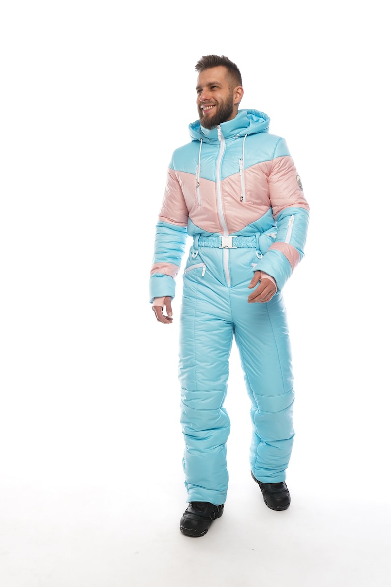 Op de afbeelding: Een lichtblauw en roze skipak met een capuchon en een witte rits. Het pak heeft een riem en is gemaakt van een gewatteerd materiaal.
