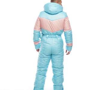 Op de afbeelding: Een lichtblauw skibroek met een roze paneel op de rug. Het pak heeft een capuchon en een ritssluiting.