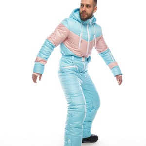 Op de afbeelding: Een lichtblauw en roze skipak met een capuchon. Het pak heeft een ritssluiting en een riem. De persoon draagt zwarte laarzen.
