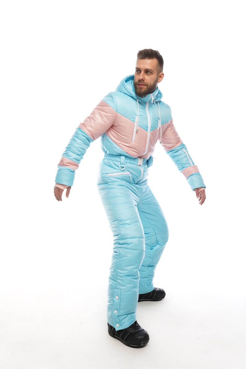 Op de afbeelding: Een lichtblauw en roze skipak met een capuchon. Het pak heeft een ritssluiting en een riem. De persoon draagt zwarte laarzen.