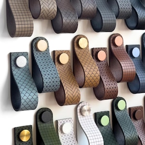Puede incluir: Una colección de tiradores de cuero para armarios en varios colores y patrones. Cada tirador presenta un diseño de bucle con un botón metálico decorativo. Los colores incluyen tonos de marrón, azul, verde y blanco. Los tiradores están montados sobre una superficie blanca.
