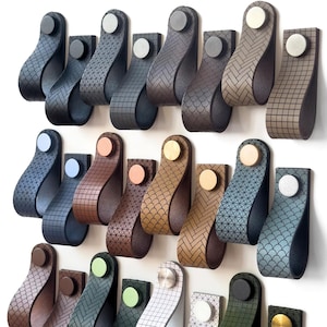 May include: A selection of leather cabinet pulls in a variety of colours and patterns. The pulls are mounted on a white wall and feature different metal accents, including silver, gold, and copper.