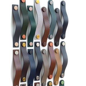Puede incluir: Una colección de tiradores decorativos de cuero para armarios en varios colores, incluyendo negro, marrón y azul. Cada tirador presenta un diseño curvo y está sujeto con herrajes metálicos. Los tiradores tienen diferentes texturas, como perforadas y estampadas.