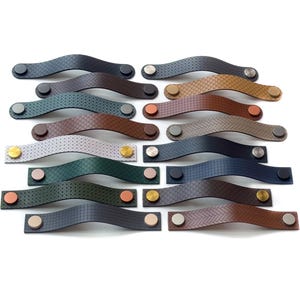 Könnte beinhalten: Eine Sammlung von Leder-Schrankgriffen in verschiedenen Farben, darunter Schwarz, Braun und Blau. Jeder Griff hat ein geschwungenes Design mit dekorativen Nähten und ist mit Metallnieten befestigt. Die Griffe sind auf weißem Hintergrund angeordnet.