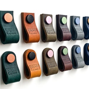 May include: A collection of 15 leather drawer pulls in various colours, including black, brown, green, blue, and grey. Each pull features a round knob and the text "pipes at home".
