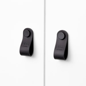 Könnte beinhalten: Zwei schwarze Leder-Schrankgriffe mit einer schwarzen Metallschraube. Die Griffe haben den Text "PIPA  AT HOME" geprägt.