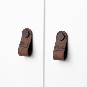 Könnte beinhalten: Zwei braune Leder-Schrankgriffe mit einer schwarzen Metallschraube. Die Griffe haben den Text "pipes at home" geprägt.