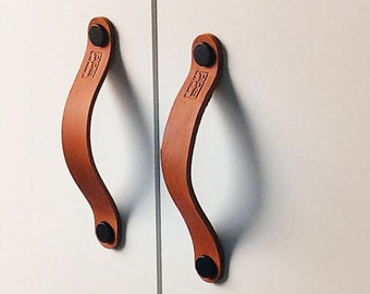 Può includere: Due maniglie per armadi in pelle marrone con accenti in metallo nero. Le maniglie hanno la forma di una 'D' arrotondata e un piccolo logo rettangolare sulla pelle.