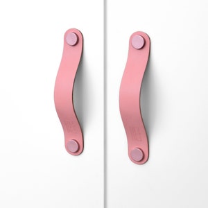 Peut inclure: Deux poignées de tiroir en cuir rose avec une forme rectangulaire arrondie et un petit bouton rond à chaque extrémité.