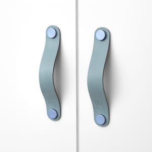 Puede incluir: Dos tiradores de armario de cuero azul claro con detalles metálicos plateados. Los tiradores están fijados a una superficie blanca.