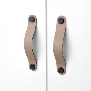 Könnte beinhalten: Zwei taupefarbene Leder-Schrankgriffe mit schwarzen kreisförmigen Befestigungen. Die gebogenen Griffe sind an einer weißen Schranktür befestigt. Die Griffe haben ein minimalistisches Design und eignen sich für moderne Wohnkultur.