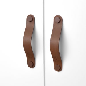 Peut inclure: Deux poignées de tiroir en cuir marron avec une forme arrondie et incurvée. Les poignées ont un petit bouton rond marron à chaque extrémité.