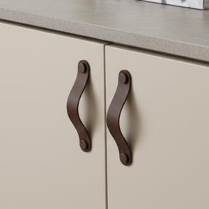 Op de afbeelding: Close-up van twee kastdeuren met donkerbruine leren handgrepen. De handgrepen zijn gebogen en bevestigd met klinknagels. De kastdeuren zijn lichtbeige van kleur en het aanrechtblad erboven is lichtgrijs.