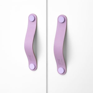 Könnte beinhalten: Zwei lilafarbene Leder-Schrankgriffe mit jeweils einem runden, hellvioletten Knopf an den Enden.