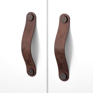 May include: Two brown leather cabinet pulls with metal rivets. The pulls are curved and have the text "PIGGS" printed on them.