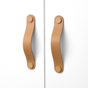 May include: Two tan leather cabinet pulls with brass accents. The pulls are attached to a white surface.