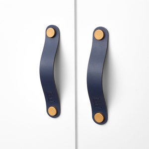 May include: Two blue leather cabinet pulls with gold metal accents. The pulls are attached to a white surface.