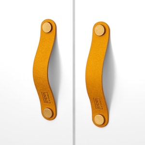 May include: Two mustard yellow leather cabinet pulls with brass accents. The pulls have a curved shape and are branded with "PIPES" in a black rectangle.