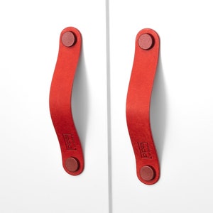 Könnte beinhalten: Zwei rote Leder-Schrankgriffe mit einer geschwungenen Form und einem runden Metallknopf an jedem Ende. Die Griffe haben den Text "PIPES" aufgedruckt.