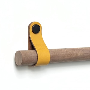 Puede incluir: Una barra de madera con una correa de cuero amarilla y un tornillo de metal negro. La correa está unida a la barra y tiene la palabra "BOHO" grabada en relieve.