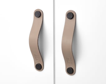 Può includere: Due maniglie per armadi in pelle color talpa con elementi di fissaggio circolari neri. Le maniglie curve sono attaccate a un'anta bianca dell'armadio. Le maniglie hanno un design minimalista e sono adatte all'arredamento moderno.
