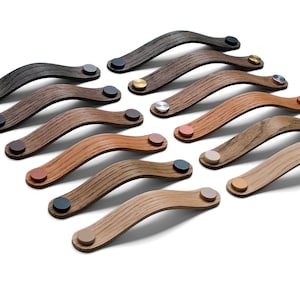 May include: A collection of wooden cabinet pulls in various shades of brown, from dark to light. Each pull has a curved design and is affixed with a small, circular metal accent in colors like silver, gold, and copper. The pulls are arranged on a white background.