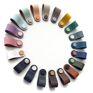 Könnte beinhalten: Ein Set mit 18 Leder-Schubladengriffen in verschiedenen Farben, darunter Rosa, Blau, Grün, Braun und Gelb. Jeder Griff hat einen Metallknopf und eine einzigartige Farbe.