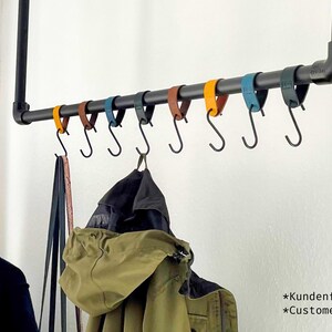 May include: A black metal pipe clothing rack with a horizontal bar holding several S-hooks. The hooks are attached to colorful leather straps in shades of yellow, brown, blue, and black. Coats and other garments hang from the hooks.
