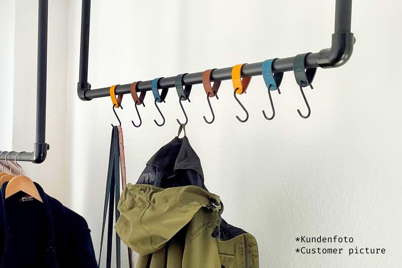 May include: A black metal pipe clothing rack with a horizontal bar holding several S-hooks. The hooks are attached to colorful leather straps in shades of yellow, brown, blue, and black. Coats and other garments hang from the hooks.