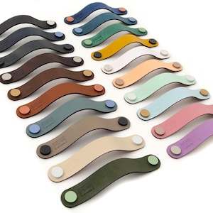 May include: A collection of 16 leather cabinet pulls in various colours, including blue, green, brown, orange, pink, and purple. Each pull has a curved shape and a small metal knob on each end.