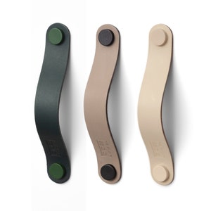 May include: Three leather cabinet pulls in dark green, tan, and beige, each with a curved design. The pulls have a circular fastener at each end, with a green, black, and beige color scheme. The pulls are mounted on a white surface.
