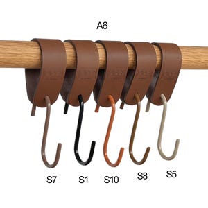 May include: Six brown leather hanging hooks with metal hooks in various colors, including black, orange, and beige, are attached to a wooden rod. The hooks are labeled with letters and numbers.